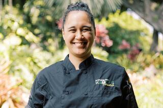 A smiling chef in a black uniform stands outdoors, surrounded by lush greenery and vibrant flowers, exuding warmth and passion for cooking.