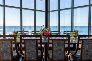 A beautifully set dining table with floral arrangements overlooks a stunning ocean view through large glass windows.