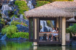A couple relaxes in a tropical setting with a thatched-roof pavilion, surrounded by lush greenery and soothing waterfalls.