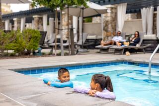 Two children chat playfully in a pool while adults relax nearby on loungers, enjoying a sunny day at a sophisticated outdoor setting.