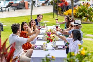 A group of friends joyfully toasts with wine glasses at a festive outdoor gathering, surrounded by vibrant flowers and greenery.