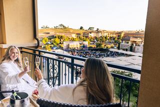 Two women in bathrobes toast with champagne on a balcony, overlooking an event area filled with an audience and scenic surroundings.