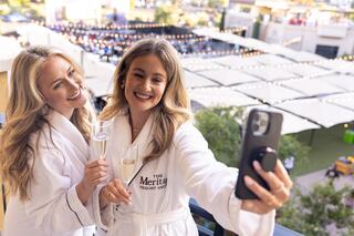 Two women in robes toast with champagne while taking a selfie on a balcony, enjoying a sunny day and festive atmosphere.