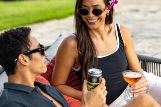 A man and woman relax outdoors, enjoying drinks and sharing smiles on a sunny day, surrounded by vibrant greenery and cozy seating.